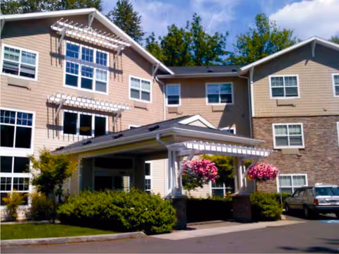 Exterior view of a multi-story senior living facility building with beige siding and stone accents. The entrance features a covered driveway with white columns and hanging flower baskets. There are bushes and trees around the building and a parked car is visible on the right side.
