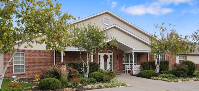 Front exterior view of a single-story brick building with a gabled roof, white trim, and a covered entrance. The entrance has a white door decorated with a wreath and flanked by windows. There are small trees, bushes, and landscaped plants along the walkway leading to the door under a partly cloudy sky.