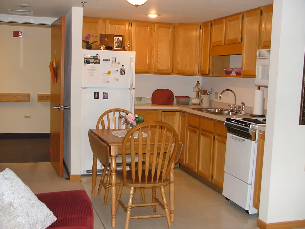 Interior view of a kitchen in a retirement community featuring wooden cabinets, a white refrigerator with magnets and photos, a white stove, a microwave, a double sink, and a small wooden dining table with two chairs and a flower centerpiece.
