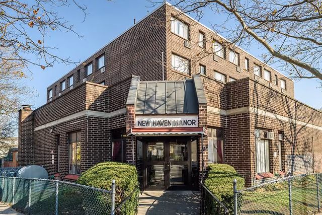 Exterior view of a multi-story brick building with a sign above the entrance that reads 'New Haven Manor'. The entrance has glass double doors and is flanked by bushes and a chain-link fence. Leafless tree branches are visible against a clear blue sky.