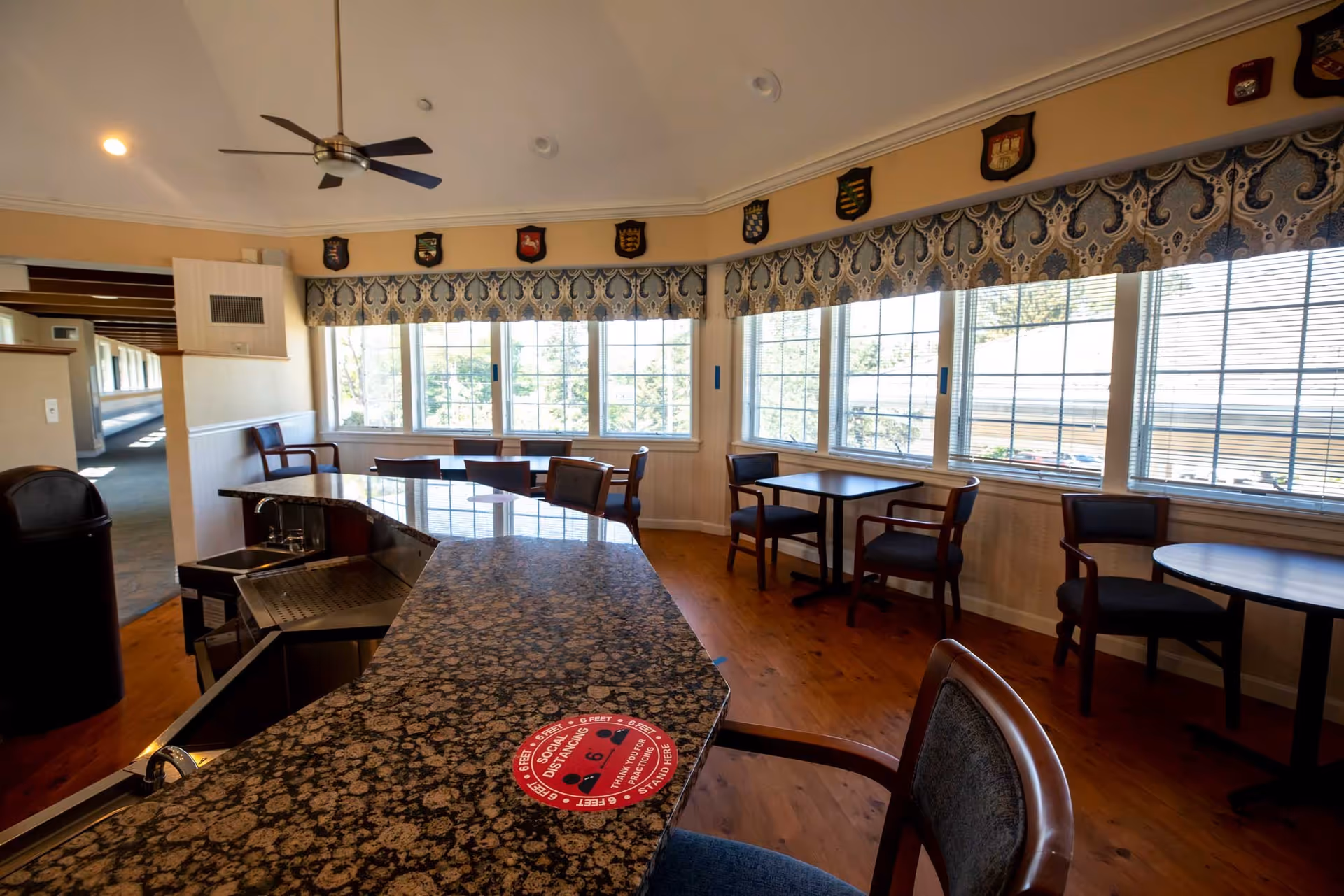 Interior view of a common area in Edelweiss Village featuring a curved granite countertop with a sink, several wooden tables and chairs near large windows with patterned valances, a ceiling fan, and decorative shields mounted on the wall above the windows.