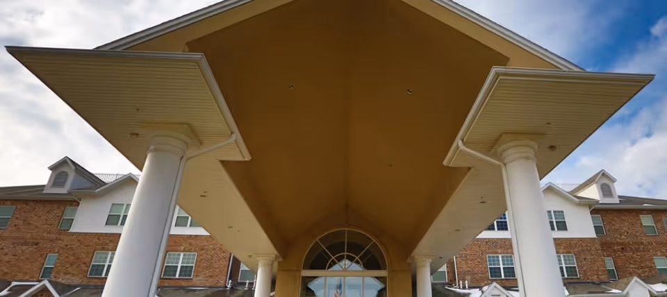 Entrance of a multi-story brick and white siding building with large white columns supporting a peaked roof over the entryway under a partly cloudy sky.
