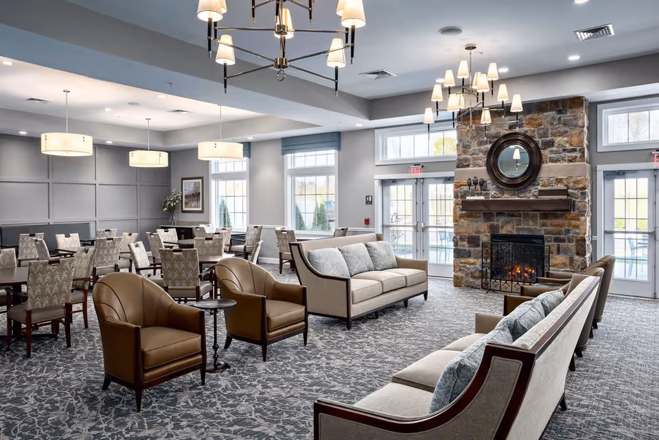 A spacious and well-lit senior living common area featuring comfortable beige sofas with blue cushions, brown leather armchairs, and multiple dining tables with patterned chairs. The room has large windows allowing natural light, a stone fireplace with a round mirror above it, and modern ceiling lights.