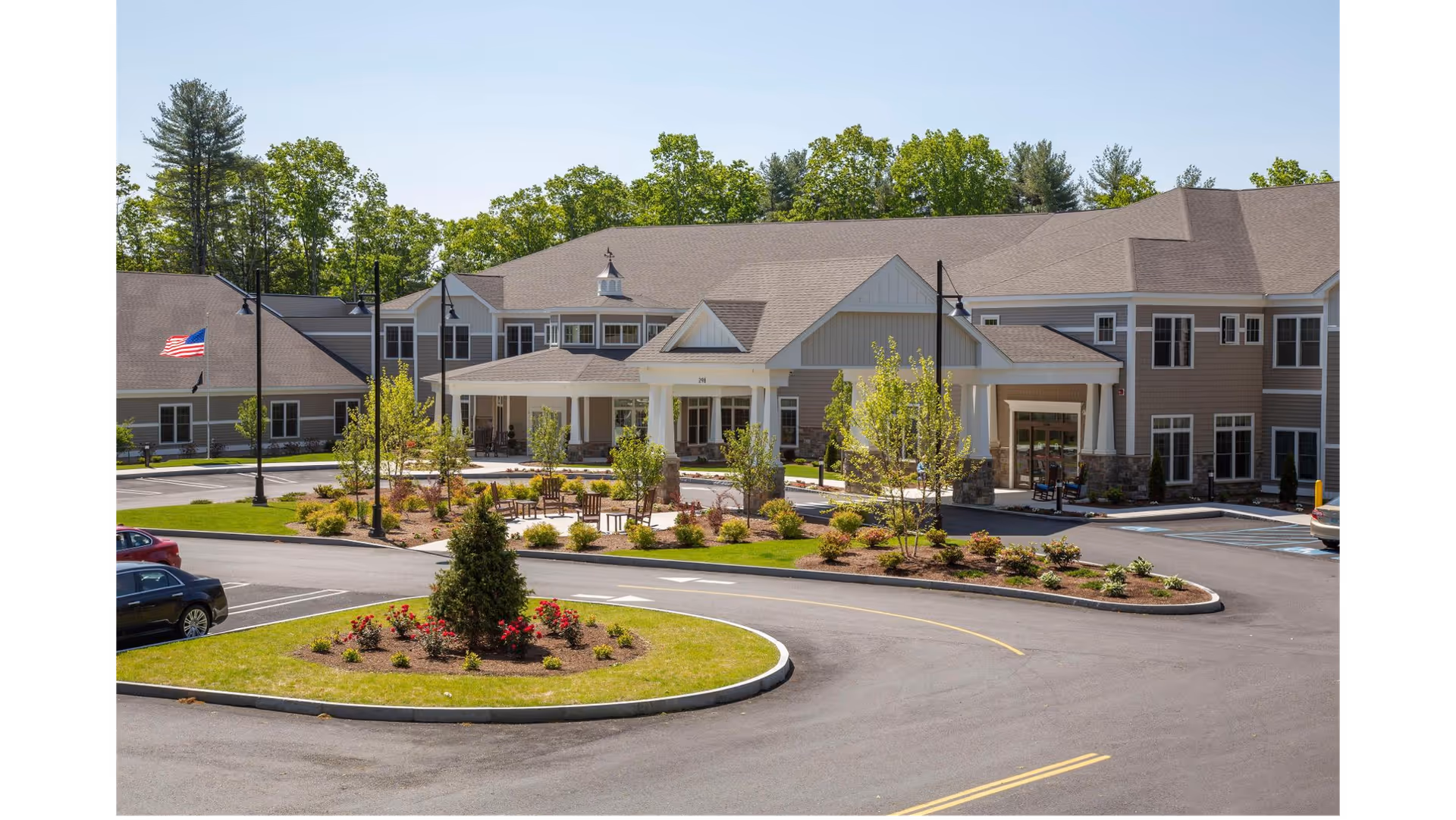 Exterior view of Cornerstone at Hampton senior living facility showing a large two-story building with a covered entrance, landscaped garden areas with shrubs and flowers, a circular driveway, parked cars, and an American flag on a flagpole. Trees are visible in the background under a clear sky.