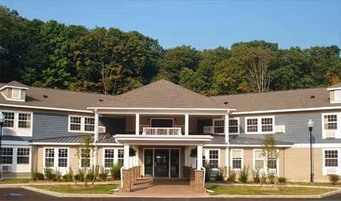 Front exterior view of a two-story senior living facility building with a covered entrance, multiple windows, and surrounded by greenery and trees.