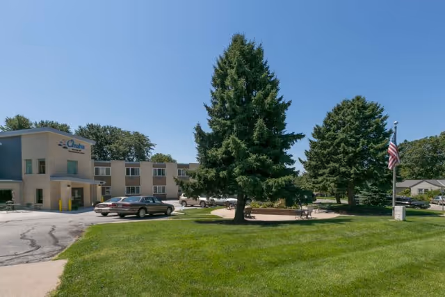 Exterior view of Omaha Nursing And Rehabilitation Center building with a parking area, green lawn, tall trees, and an American flag on a flagpole under a clear blue sky.