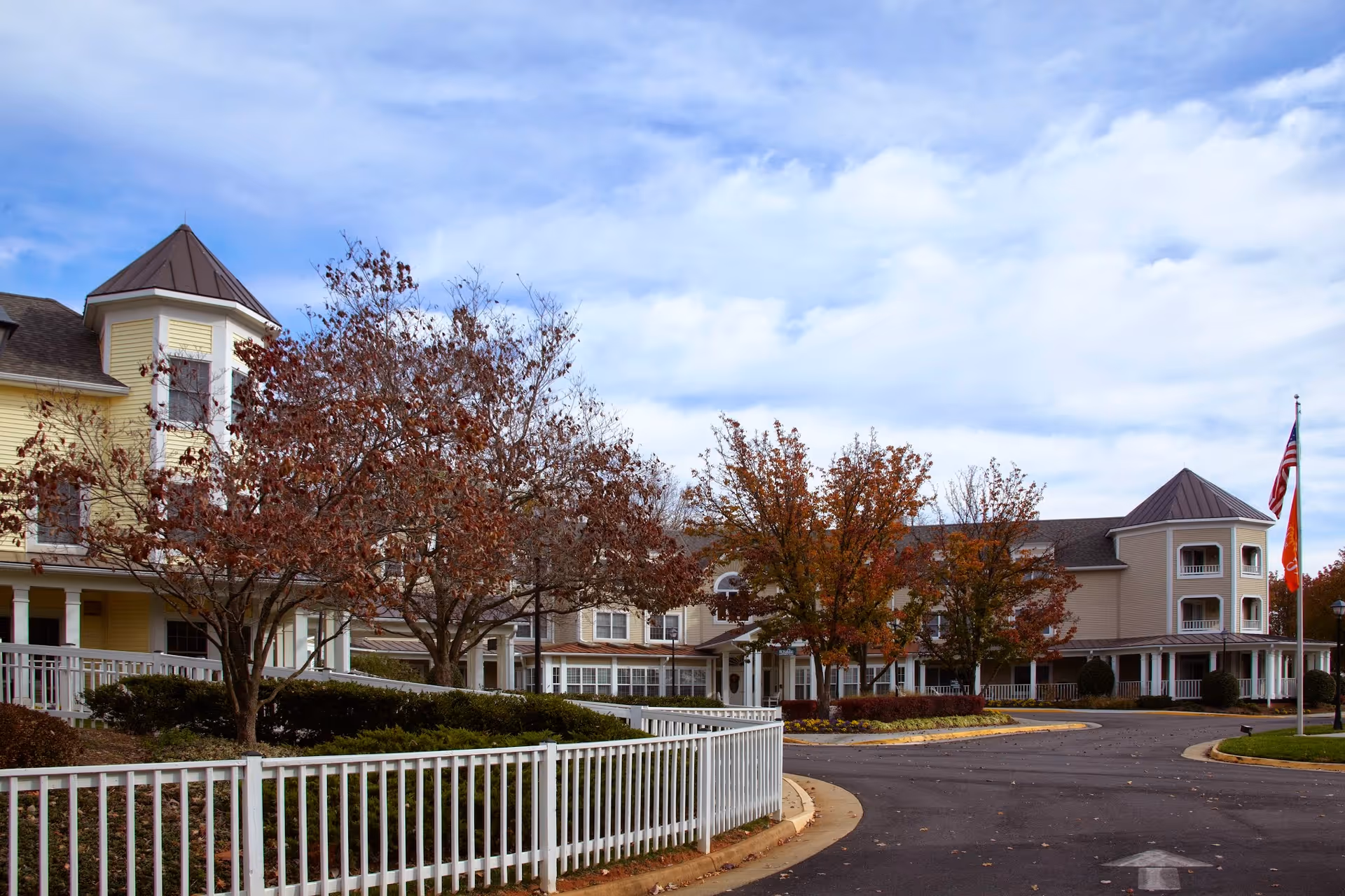 Exterior view of a senior living facility with a curved driveway, white fencing, and trees with autumn foliage. The building has multiple stories with balconies and a turret-like structure. An American flag is visible on the right side of the image under a partly cloudy sky.