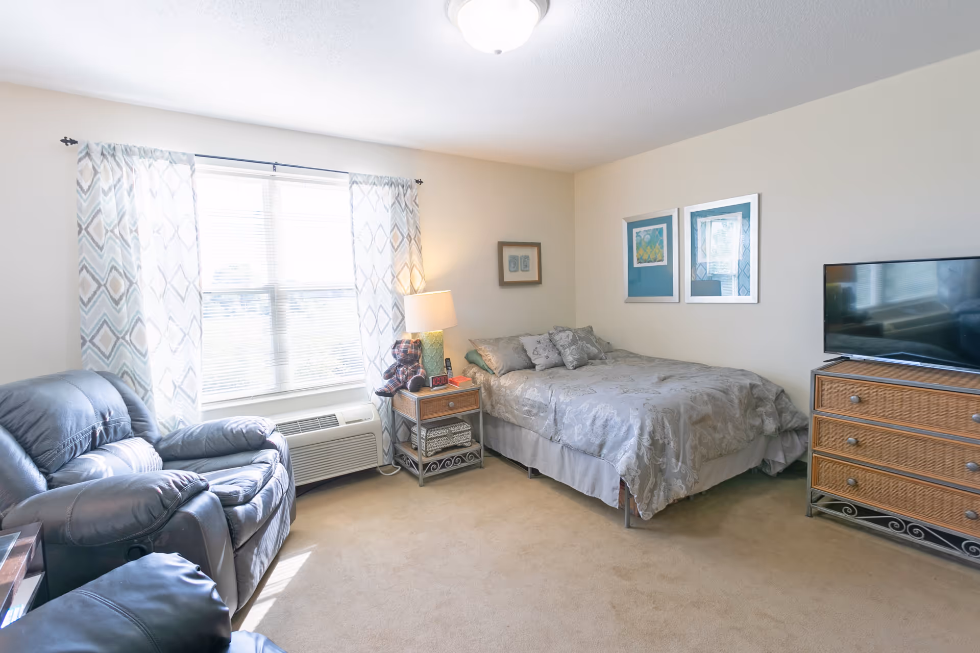 A bright and cozy bedroom with a bed covered in gray patterned bedding, a wooden nightstand with a lamp and a teddy bear, a window with patterned curtains, a leather recliner chair, and a wooden dresser with a flat-screen TV on top.