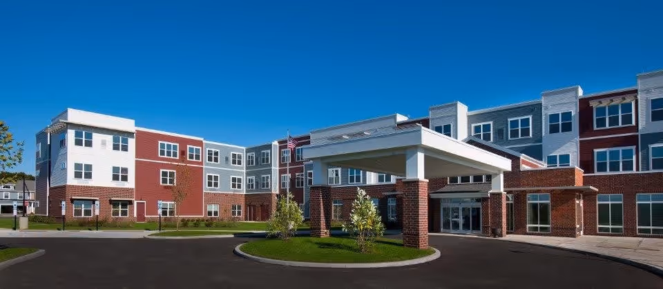 Exterior view of a modern three-story senior living facility building with a covered entrance, brick and siding facade, multiple windows, and a clear blue sky.