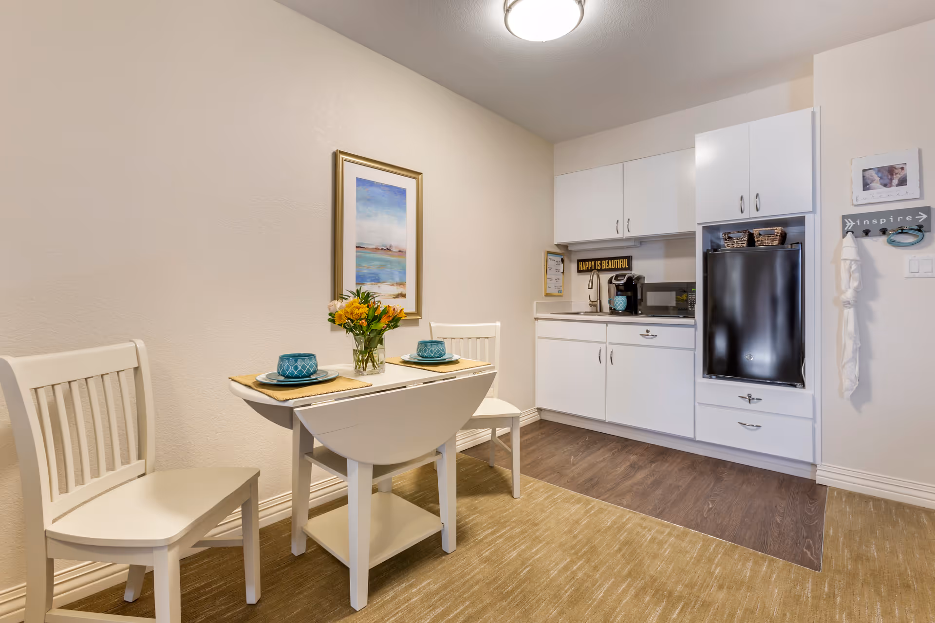 A small kitchenette and dining area with a white drop-leaf table set for two with teal cups and plates, two white chairs, a vase with flowers, white cabinets, a black mini refrigerator, a microwave, and a coffee maker. The wall has a framed abstract painting and decorative signs including one that says 'HAPPY IS BEAUTIFUL'.