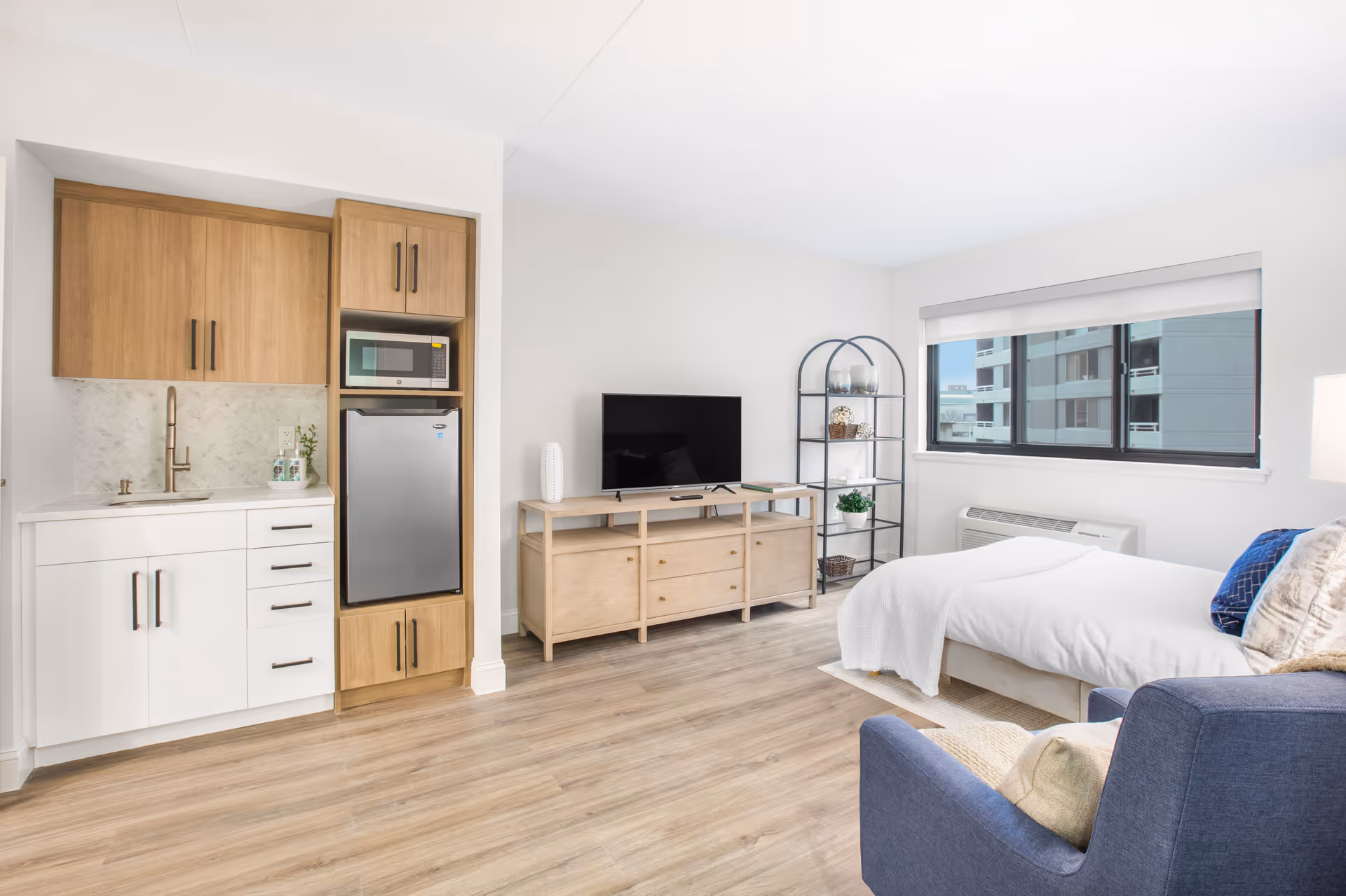 A bright and modern studio apartment featuring a small kitchenette with wooden cabinets, a microwave, and a mini refrigerator on the left. In the center, there is a wooden TV stand with a flat-screen television and decorative items. To the right, a bed with white bedding is positioned near a large window with a view of nearby buildings. A blue armchair with cushions is in the foreground, and a black metal shelving unit with plants and decor is next to the window. The room has light wood flooring and white walls.