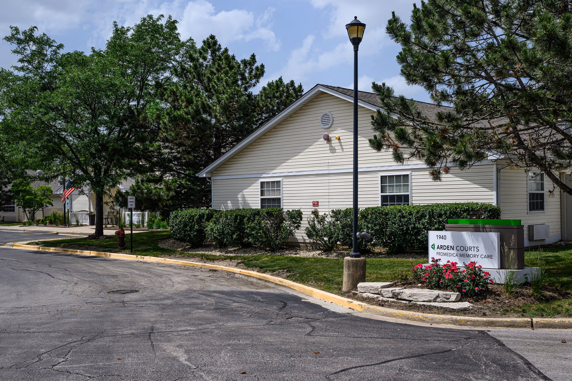 Exterior view of Arden Courts - ProMedica Memory Care Community building with beige siding, surrounded by green trees and bushes. A street lamp stands near a sign that reads '1940 Arden Courts ProMedica Memory Care' with flowers planted around the sign. The sky is partly cloudy.