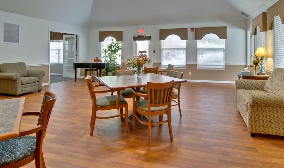 A spacious, well-lit common area with wooden flooring featuring a square table with four wooden chairs around it. There are two armchairs and a sofa along the walls, a lamp on a side table, and a piano near the back wall with large windows covered by blinds and valances.