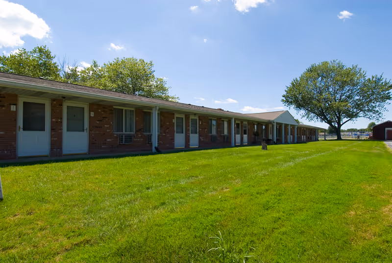 Single-story brick building with multiple white doors and windows, situated next to a large green lawn under a blue sky with some clouds. There are trees in the background and a small shed visible to the right.