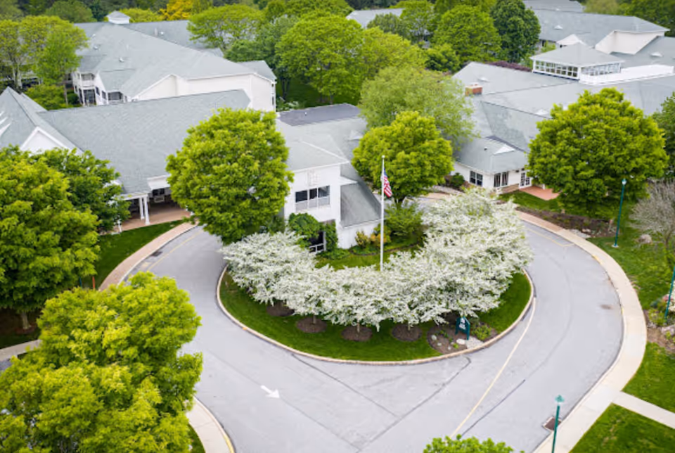 Aerial view of White Horse Village showing a circular driveway with a landscaped island featuring white flowering bushes and an American flag. Surrounding the driveway are multiple buildings with gray roofs and lush green trees.
