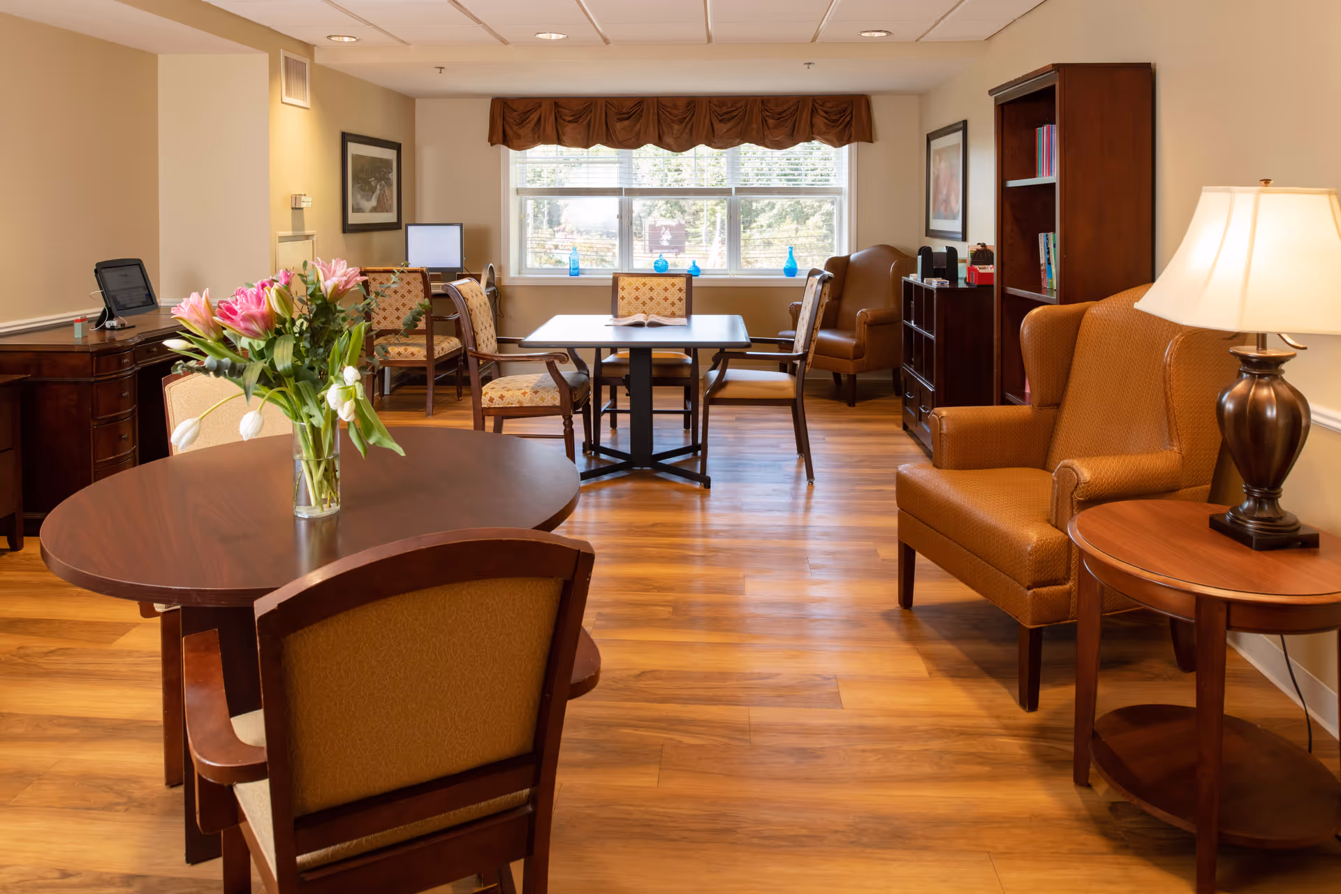A well-lit senior living common area with wooden floors, featuring a round table with a vase of flowers, several upholstered chairs, a square table with chairs near a window, a bookshelf, and a table lamp on a side table.