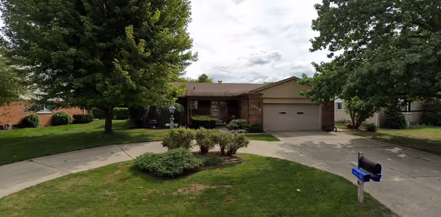 Single-story brick house with a two-car garage, surrounded by green trees and a well-maintained lawn. A curved driveway leads to the garage, and a blue mailbox is visible near the street.