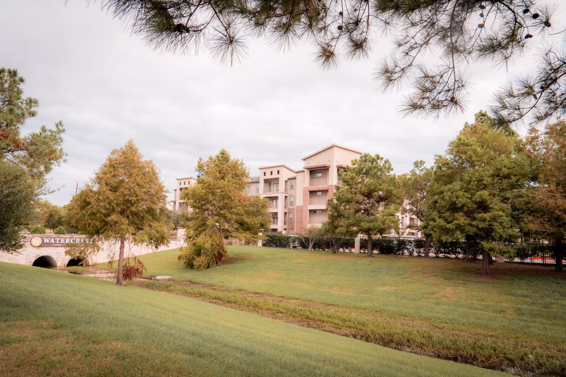 Exterior view of Watercrest at Shadow Creek Ranch facility showing a multi-story building surrounded by trees and a well-maintained grassy area with a small waterway and a stone bridge with the Watercrest sign.