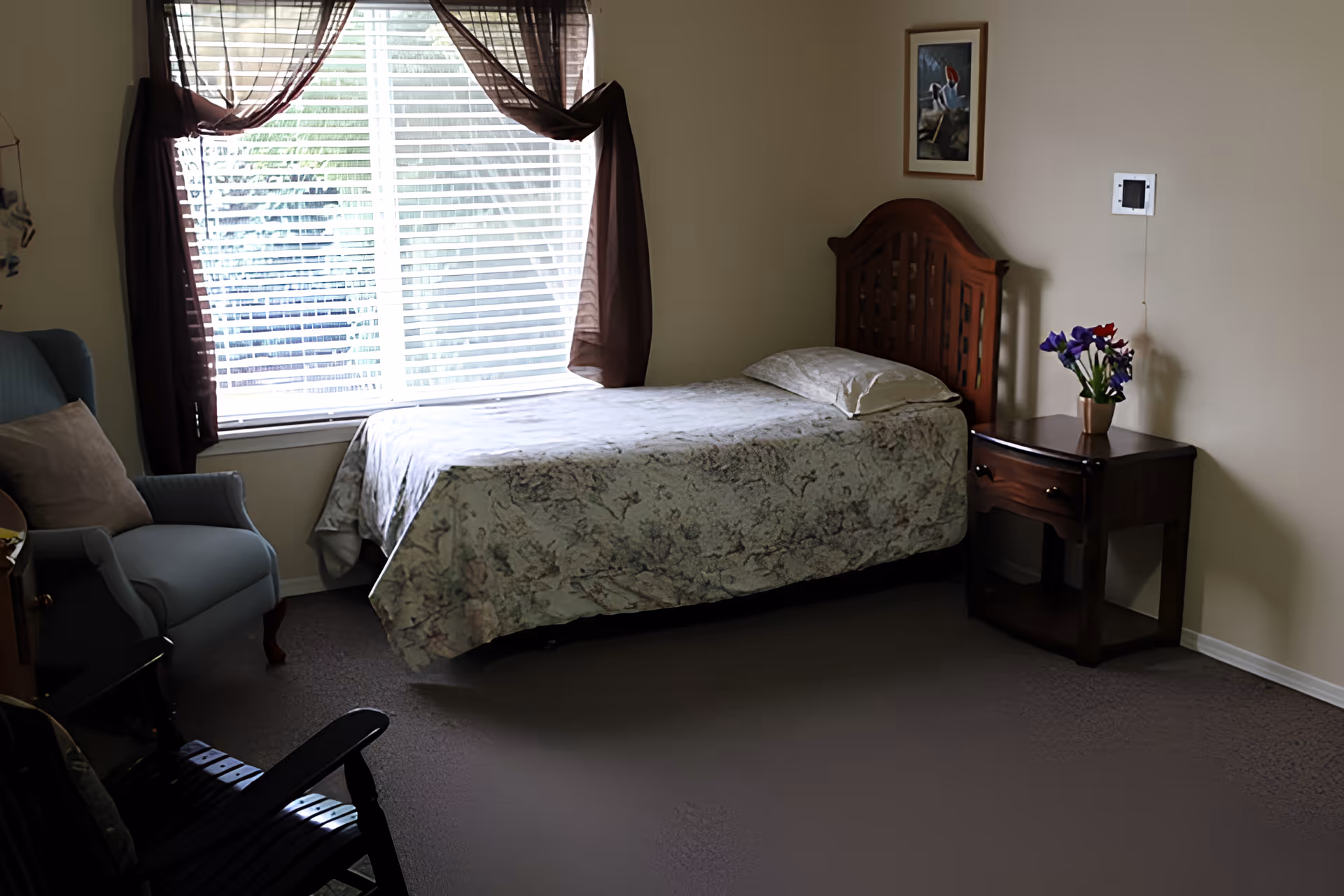 A small bedroom with a single bed covered in a floral bedspread positioned against a wall. Next to the bed is a wooden nightstand with a vase of purple and red flowers. A framed picture hangs on the wall above the nightstand. A window with white blinds and sheer brown curtains lets in natural light. There is a blue armchair with a beige pillow and a black rocking chair in the room.