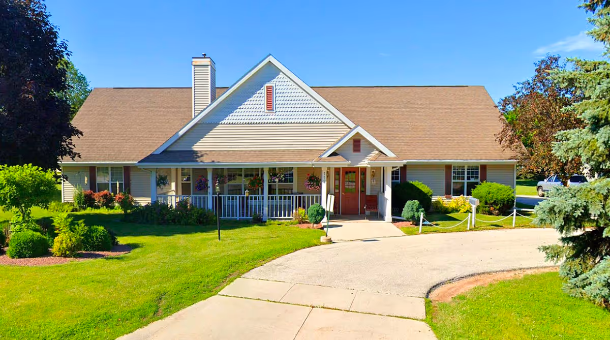 Front exterior view of a single-story senior living facility with beige siding, a brown shingled roof, a chimney, and a covered porch with hanging flower baskets. The building is surrounded by well-maintained green lawns, shrubs, and trees under a clear blue sky.