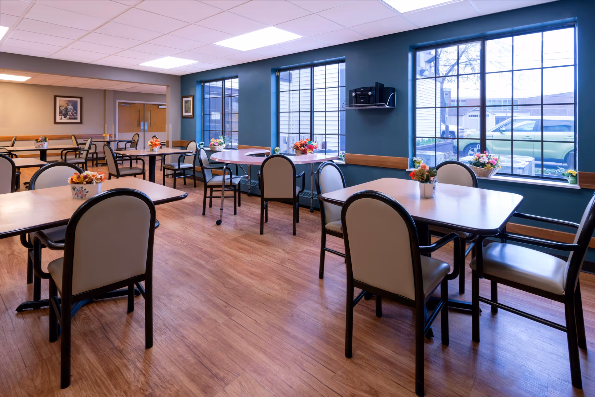 A bright dining room with multiple tables and chairs arranged neatly. Each table has a small flower arrangement. Large windows on one side let in natural light and offer a view of parked cars outside. The room has wood flooring and teal-colored walls with a white ceiling.