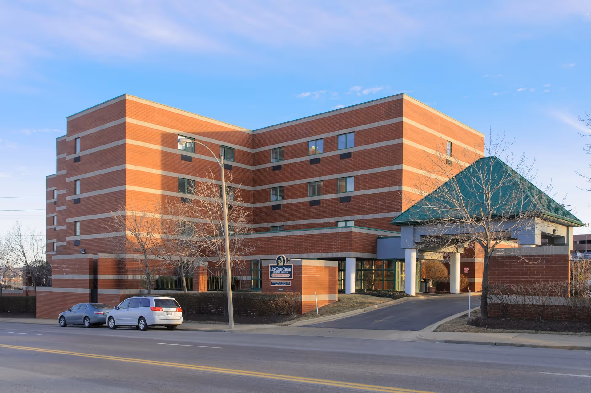 Exterior view of a multi-story brick building with horizontal light-colored stripes, identified as Life Care Center of Saint Louis. The building has several windows and a covered entrance with a green roof. There are two cars parked on the street in front of the building and leafless trees around the property under a clear blue sky.