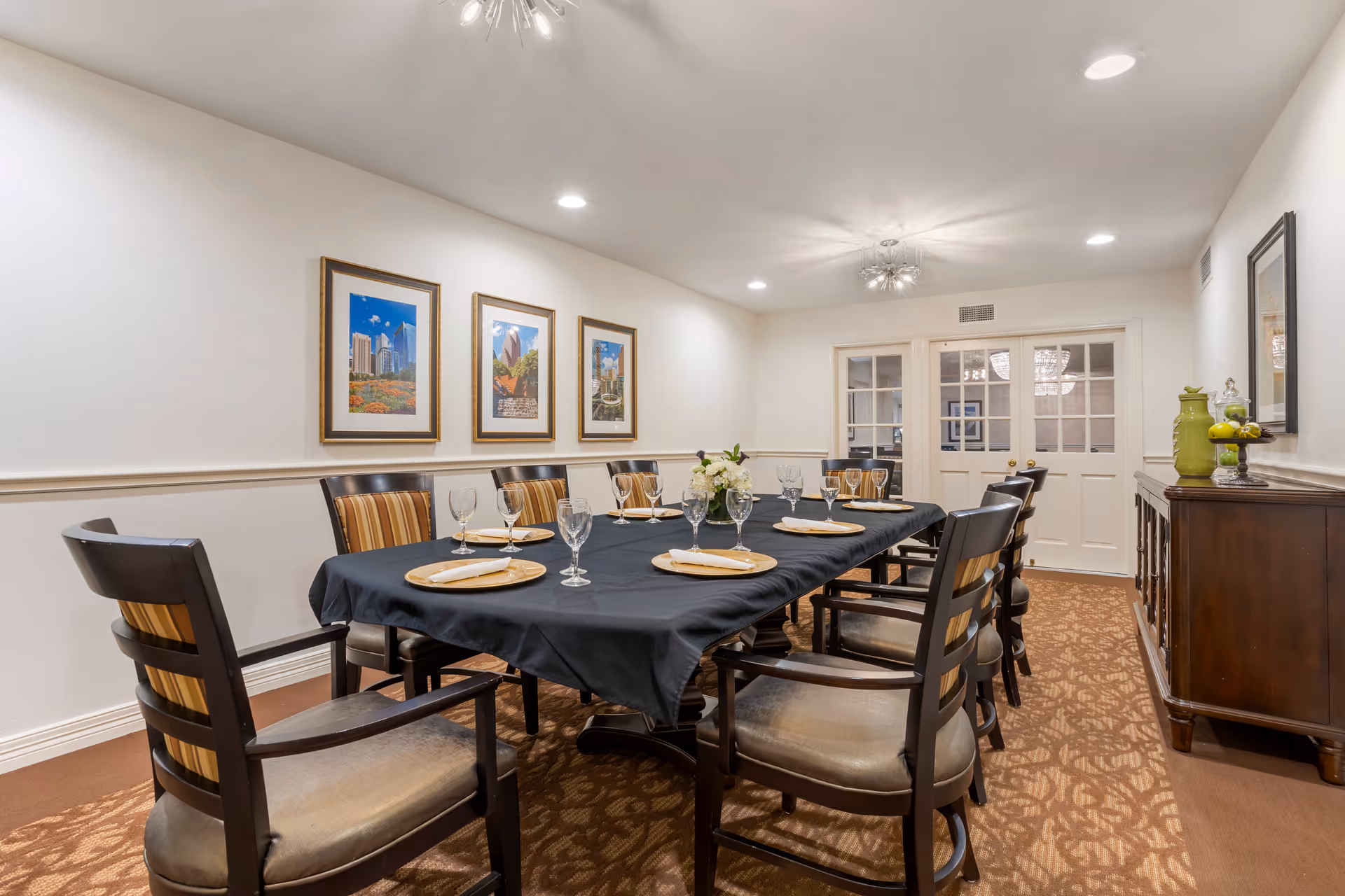A formal dining room with a long rectangular table covered with a black tablecloth. The table is set with gold plates, folded napkins, and empty wine glasses. There are ten wooden chairs with cushioned seats and striped backs around the table. The room has white walls adorned with three framed pictures of cityscapes on one side and a wooden sideboard with decorative items on the other. The floor is carpeted with a patterned design, and the ceiling has recessed lighting and a modern chandelier. Double glass-paneled doors are visible at the far end of the room.