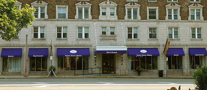 Front exterior view of a multi-story brick building with multiple windows and purple awnings. The awnings display the text 'Senior Apartments' and 'Tower Grove Manor'. The building has a central entrance with the address 2710 S. Grand above it. There are trees and a street in front of the building.