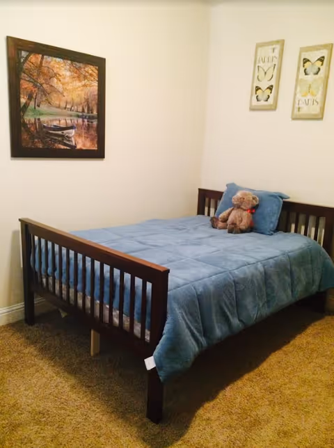 A small bedroom with a wooden bed frame, a blue comforter, and a teddy bear sitting on a blue pillow. The walls are light-colored and decorated with two butterfly-themed pictures and a framed painting of a boat on a river surrounded by autumn trees.
