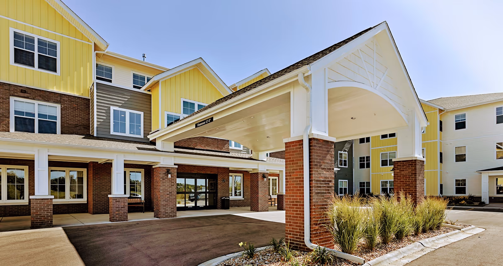 Exterior view of a senior living facility building with a covered entrance supported by brick pillars, yellow and gray siding, multiple windows, and landscaping with ornamental grasses under a clear blue sky.