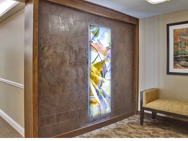 Interior hallway corner with a decorative illuminated abstract artwork mounted on a textured brown wall, a beige cushioned bench with dark wooden legs, and a framed picture on the adjacent wall.