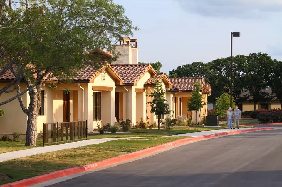 A quiet residential street in a senior living community with beige stucco buildings featuring red tile roofs. There are small trees and shrubs along the sidewalk, and two elderly people walking together on the sidewalk. The sky is partly cloudy.