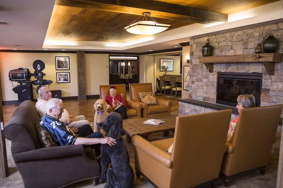 A cozy living room in a senior living facility with four elderly people sitting on sofas and armchairs around a wooden coffee table. Two dogs are also present, one black and one golden. The room features a stone fireplace with decorative items on the mantel and warm lighting from a ceiling fixture. In the background, there is a dining area with a table and chairs.