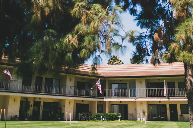 Two-story building with balconies and several American flags displayed, surrounded by tall trees and a well-maintained grassy lawn in front.