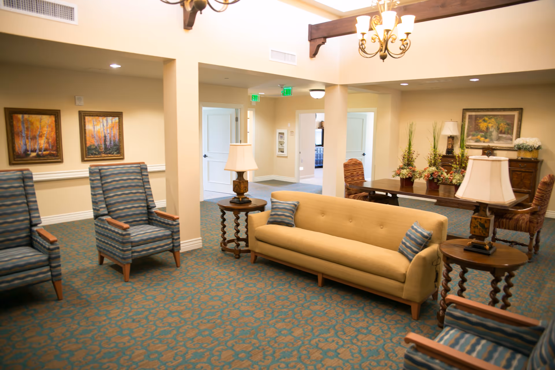 A cozy senior living facility common area with a yellow sofa, two blue patterned armchairs, wooden side tables with lamps, a wooden dining table with chairs, floral arrangements, and framed paintings on beige walls. The room has a patterned carpet and soft lighting.
