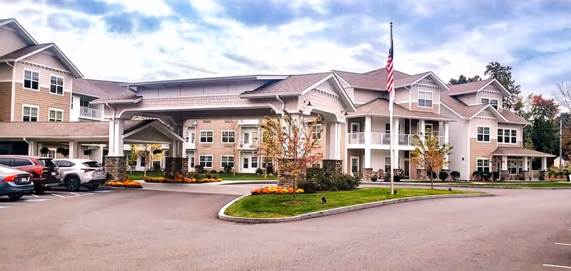 Exterior view of Kennedy Meadows Gracious Retirement Living facility showing a large multi-story building with beige siding, white trim, and a covered entrance. Several cars are parked in the parking lot, and there is a flagpole with an American flag in front of the building. The landscaping includes green grass, small trees, and flower beds.