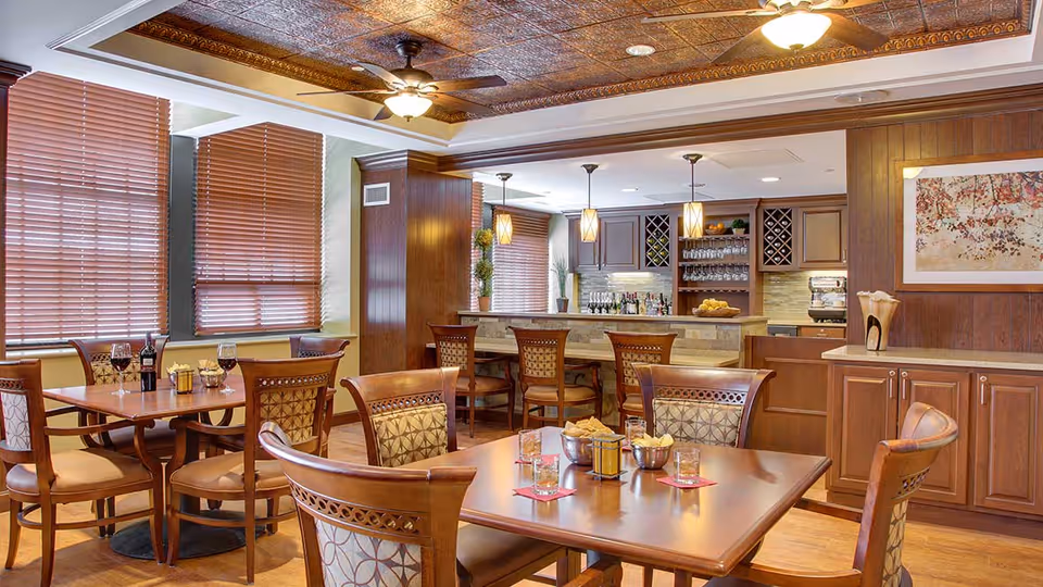 A warm and inviting dining area with wooden tables and chairs, set with glasses, bowls of snacks, and a bottle of wine. The room features large windows with wooden blinds, a ceiling fan, pendant lights above a bar counter with stools, and wooden cabinetry with a wine rack and coffee machine.