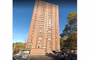Tall multi-story brick residential building under a clear blue sky with parked cars and trees around the base.