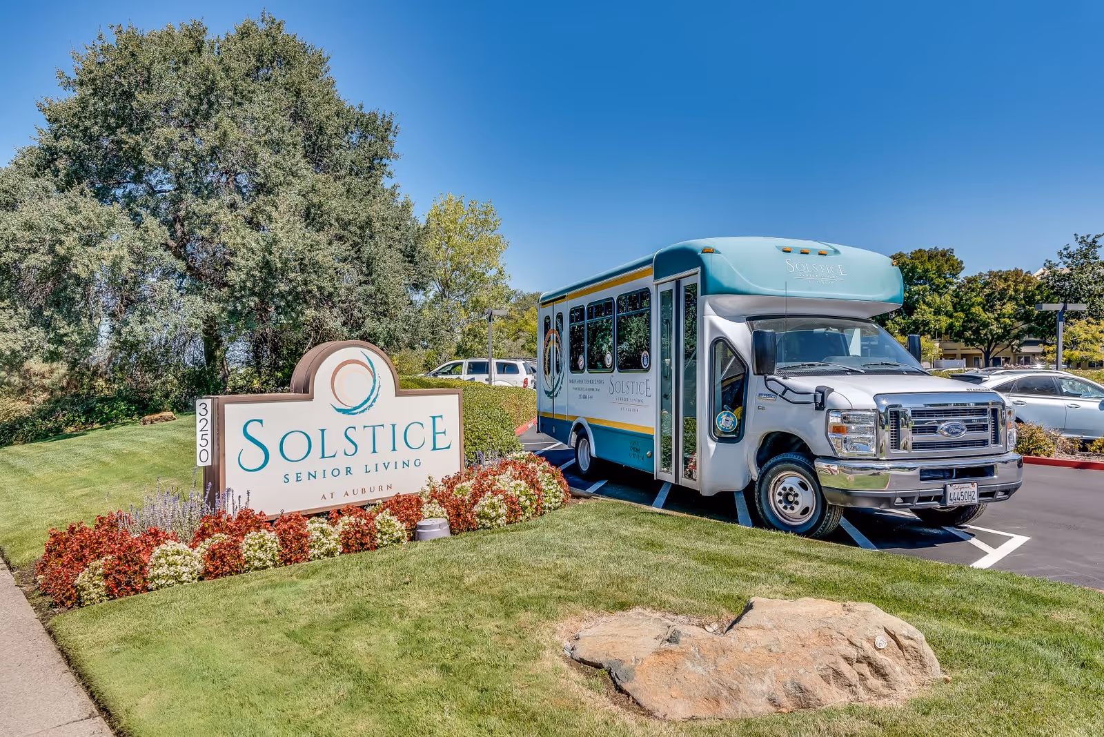 Exterior view of Solstice Senior Living at Auburn showing a large sign with the facility's name surrounded by colorful flowers and greenery. A small shuttle bus with the Solstice logo is parked nearby in a parking lot under a clear blue sky.