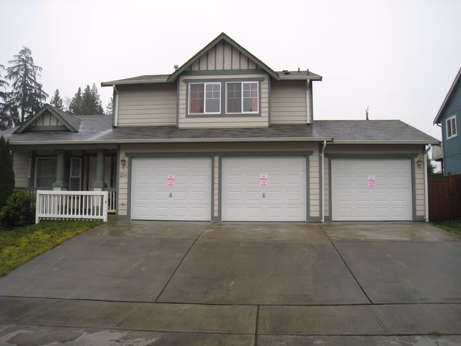 Front exterior view of a two-story residential building with three white garage doors, a small porch with white railing, and a driveway. The house is painted in light gray with green trim and has a gabled roof. There are trees and neighboring houses visible in the background under an overcast sky.