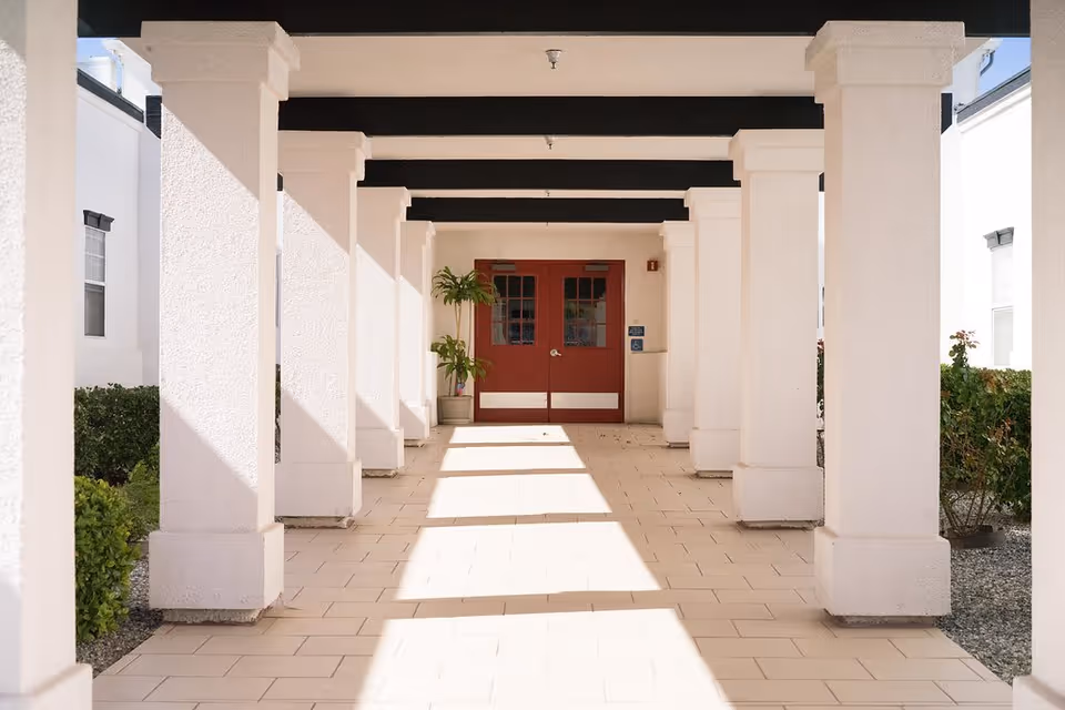 A covered outdoor walkway with white columns and a tiled floor leading to a set of double red doors. There are plants on either side of the walkway and sunlight casting shadows from the columns.