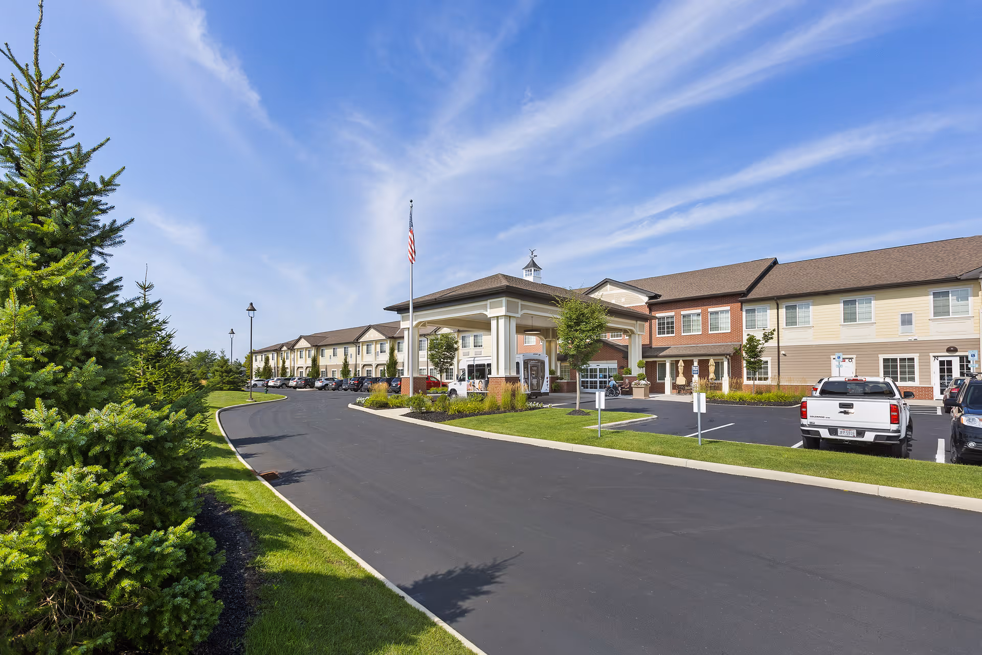 Exterior view of a senior living facility named StoryPoint Powell North with a driveway leading to the main entrance. The building is two stories tall with a covered entrance, several parked cars, green grass, and trees on the left side under a partly cloudy blue sky.