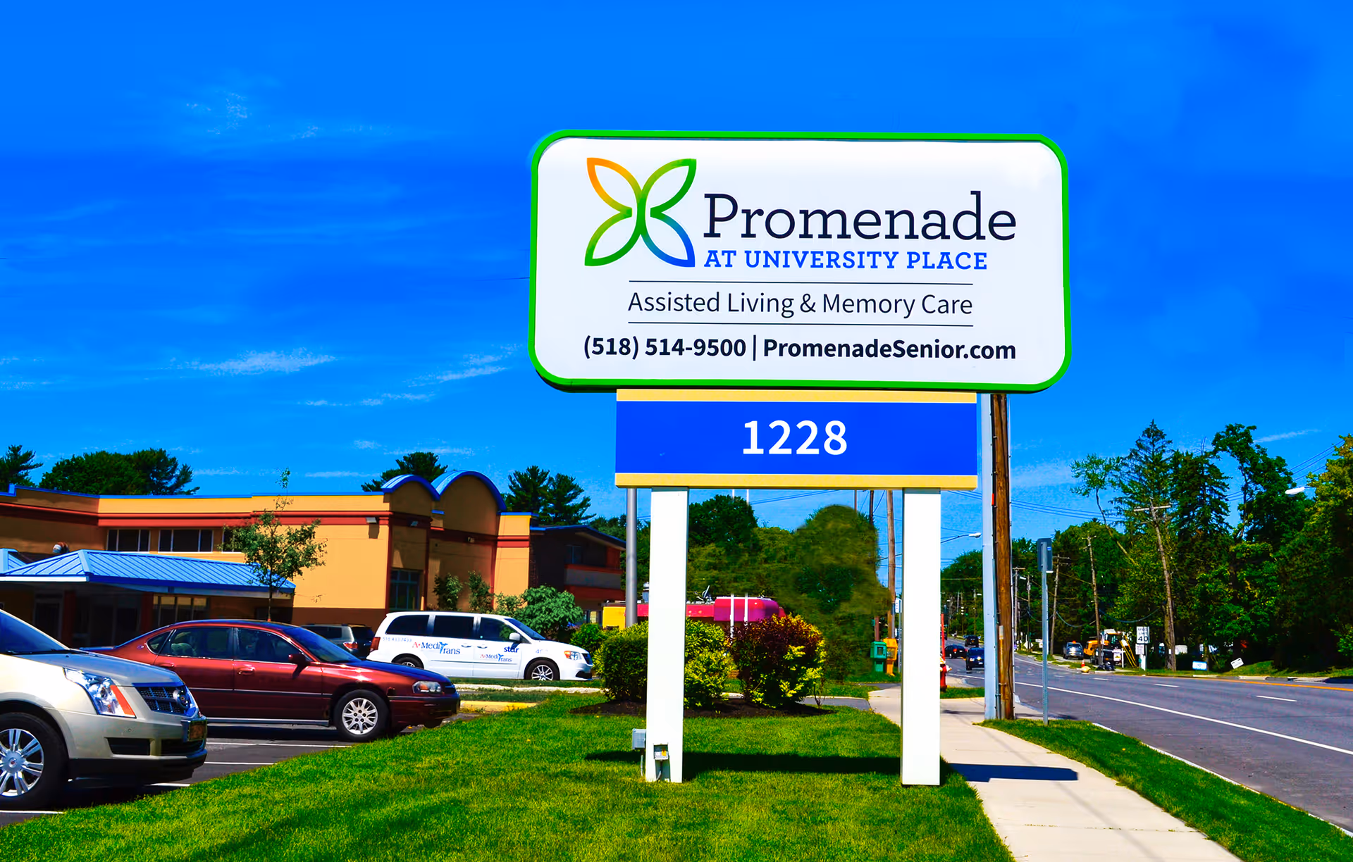 Outdoor view of a large sign for Promenade at University Place, an assisted living and memory care facility, with a blue sky background. The sign includes the phone number and website. There are cars parked on the left and a road on the right with trees and buildings in the background.