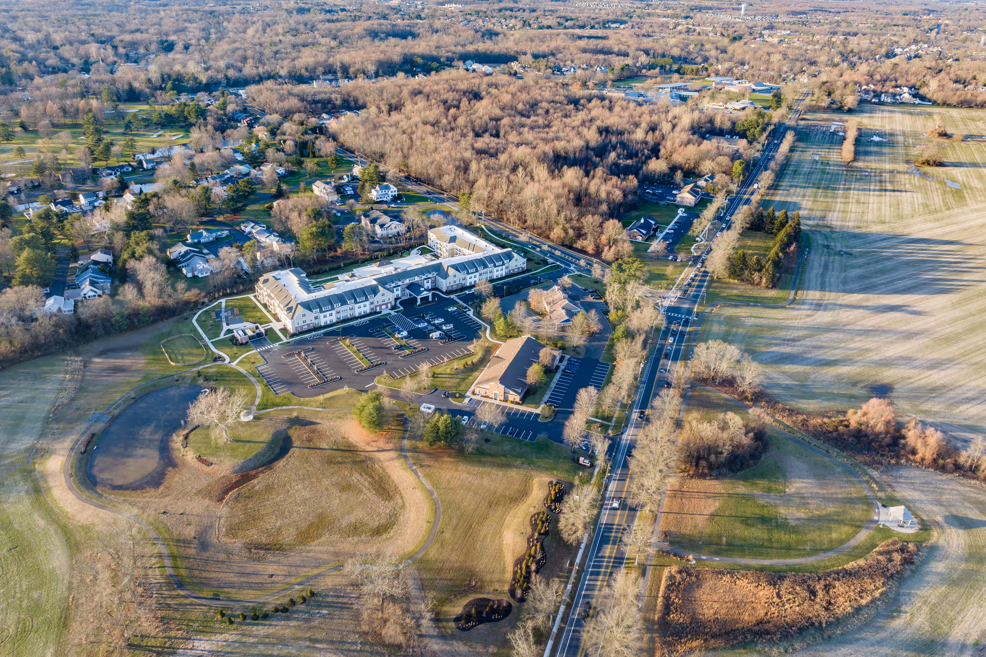 Aerial view of The Farmstead at Medford facility surrounded by open fields, trees, and a nearby residential area. The image shows the main building, parking lots, roads, and landscaped grounds under clear daylight.