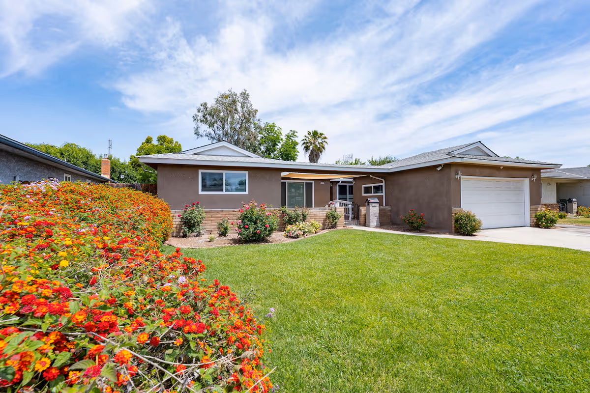 Single-story residential building with a well-maintained green lawn and colorful flowering bushes in the front yard under a partly cloudy blue sky.