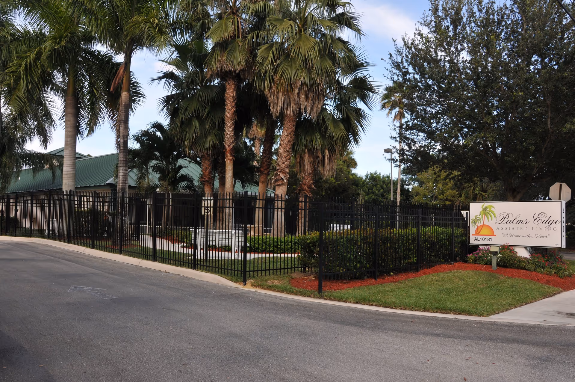 Exterior view of Palms Edge Assisted Living & Memory Care facility showing a black metal fence surrounding landscaped grounds with palm trees and other greenery. A sign with the facility name and logo is visible on the right side near the sidewalk.