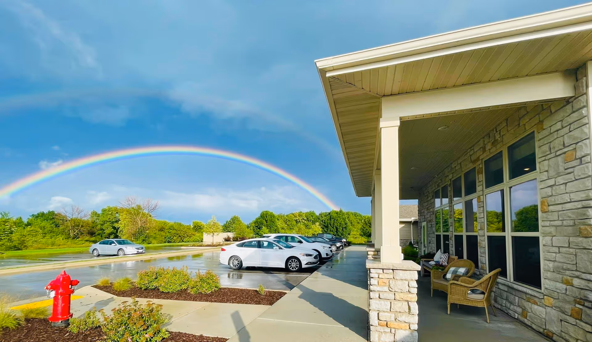 Exterior view of a senior living facility named Waterford Place showing a covered porch with wicker chairs and cushions, a parking lot with several cars, a red fire hydrant, green trees in the background, and a vibrant rainbow arching across a partly cloudy blue sky.