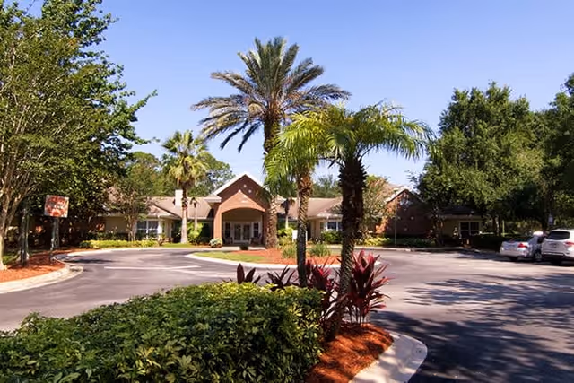 Exterior view of a senior living facility with a circular driveway, palm trees, and landscaped greenery under a clear blue sky.