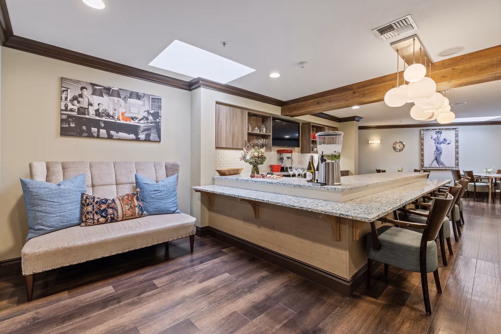 Interior view of a common area in a senior living facility featuring a long granite countertop bar with chairs, a beige cushioned bench with blue and patterned pillows, wooden flooring, and decorative artwork on the walls. The space is well-lit with ceiling lights and pendant lamps.