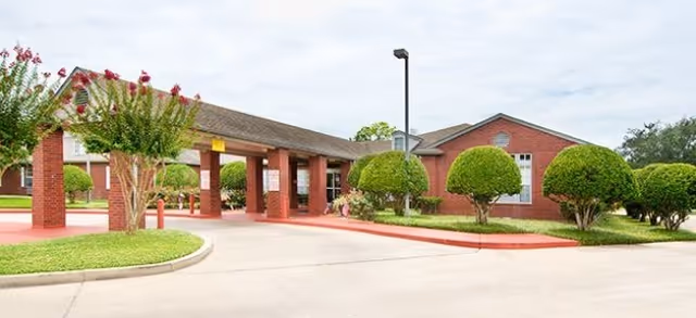 Exterior view of a senior living facility with a covered entrance supported by brick columns, neatly trimmed bushes, and a paved driveway under a partly cloudy sky.